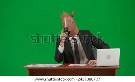 Man in business suit with horse head mask on studio green background. Businessman sits at desk, says something and points his hand to the sides. Concept of heavy office work. Royalty-Free Stock Photo #2439080797