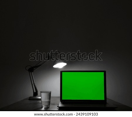 mock up. Laptop with blank green screen, and office on desk. Blank computer monitor on table with lamp light. isolated on white wall background. mockup. front view. night evening scene. glass of water