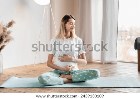 Caucasian pretty young pregnant woman holding her stomach while sitting on fitness mat in the living room. Workout, yoga and fitness at home on lockdown, pregnancy concept. Pre-natal exercises.