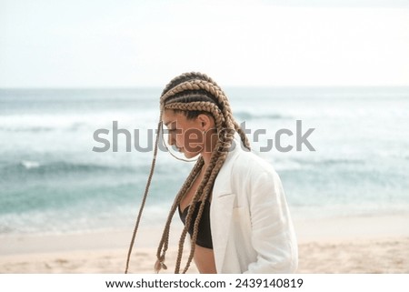 Portrait of a young Latino woman with braids on her head. She walks along the beach near the ocean in a jacket and suit. Self confidence and masculinity concept. Braiding studio. Royalty-Free Stock Photo #2439140819