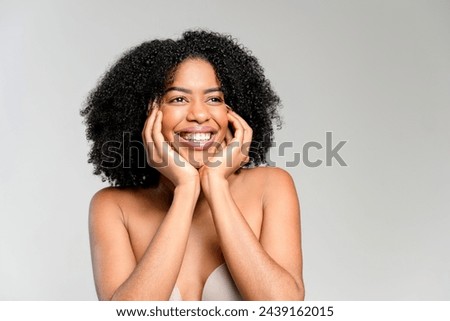 A cheerful African-American woman holds her cheeks, smiling broadly in a playful pose that embodies the joy of life against a minimalistic grey background, perfectly portraying lightheartedness. Royalty-Free Stock Photo #2439162015