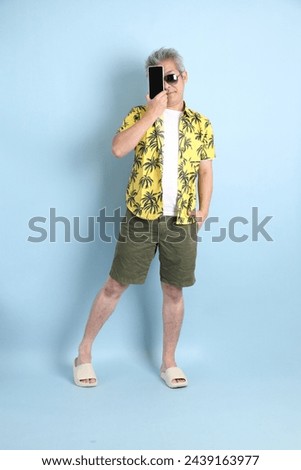 HAPPY SONGKRAN DAY. Asian energetic senior man in summer clothing and sunglasses with gesture of holding mobile phone isolated on blue background. Thai New Year's Day. Royalty-Free Stock Photo #2439163977