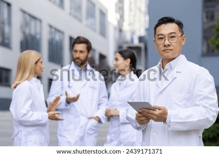 Serious chinese doctor looking at camera and holding digital tablet while standing on blurred background with colleagues. Group of medical workers in while lab coats discussing new treatment strategy. Royalty-Free Stock Photo #2439171371