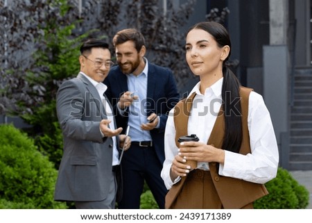 Attractive woman with coffee cup passing by two men in business suits pointing with finger and whispering behind her back. Rude males behaving unprofessional and discriminating new office worker. Royalty-Free Stock Photo #2439171389