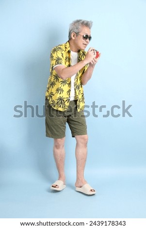 HAPPY SONGKRAN DAY. Asian energetic senior man in summer clothing and sunglasses with gesture of Holding a camera to taking a photo isolated on blue background. 