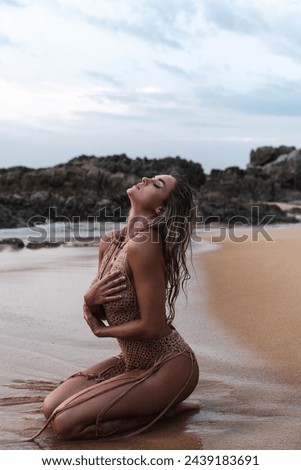 Young woman with a flawless tanned body and beautiful makeup wearing chic crochet dress as she sits gracefully on the beach.