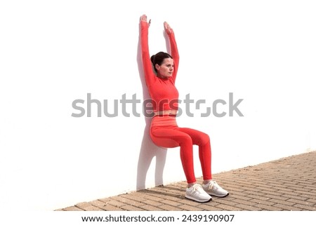 Sporty woman doing squats against a white wall with arms raised, outside in the sun. Royalty-Free Stock Photo #2439190907