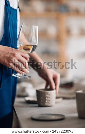Cropped picture of a pottery designer's hand holding a wineglass at ceramics studio. Wine, hobby and pottery concept. Creative craftswoman having a glass of white wine after pottery workshop in studio