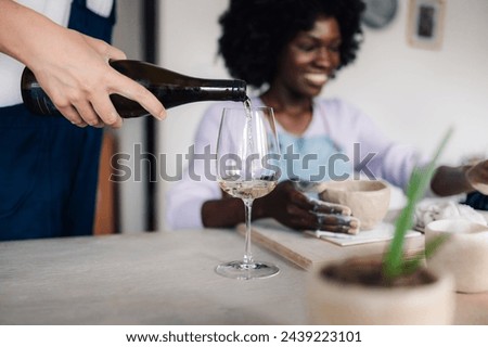 Cropped picture of a female's hand pouring wine into a wineglass on a pottery class. In a blurry background is pottery class student sitting in studio and making handmade earthenware and handcrafts.