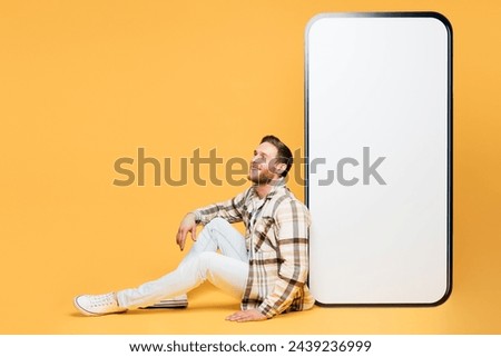Full body young man wear brown shirt casual clothes sit near big huge blank screen mobile cell phone smartphone workspace area look aside isolated on plain yellow orange background. Lifestyle concept