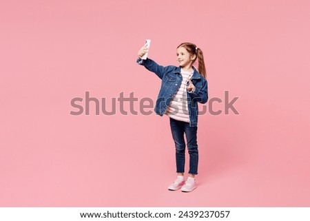 Full body little child cute kid girl 7-8 year old wear denim shirt have fun do selfie shot on mobile cell phone show v-sign isolated on plain pink background Mother's Day love family lifestyle concept
