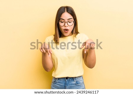 pretty hispanic woman with open mouth pointing downwards with both hands, looking shocked, amazed and surprised