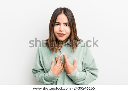pretty hispanic woman pointing to self with a confused and quizzical look, shocked and surprised to be chosen