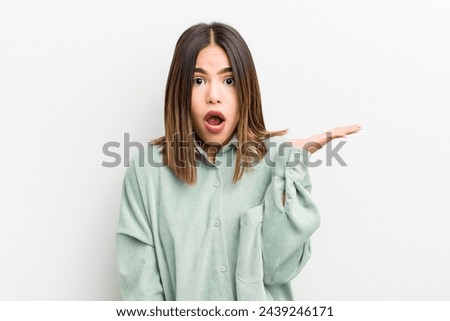 pretty hispanic woman looking surprised and shocked, with jaw dropped holding an object with an open hand on the side