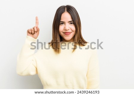 pretty hispanic woman smiling cheerfully and happily, pointing upwards with one hand to copy space