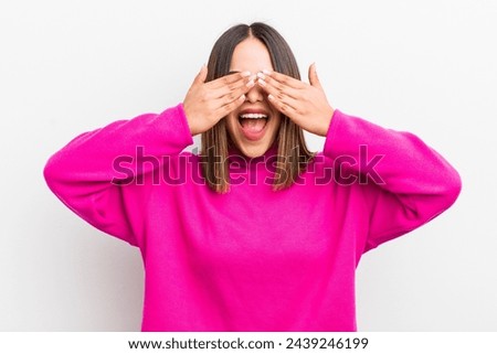 pretty hispanic woman smiling and feeling happy, covering eyes with both hands and waiting for unbelievable surprise