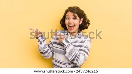 pretty hispanic woman feeling joyful and surprised, smiling with a shocked expression and pointing to the side