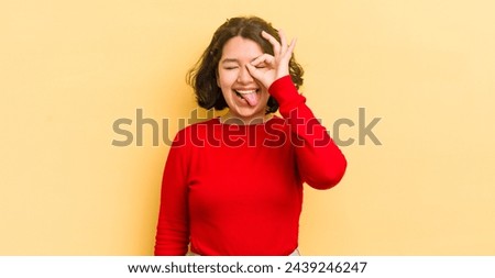 pretty hispanic woman smiling happily with funny face, joking and looking through peephole, spying on secrets