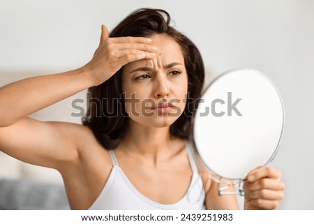 Upset millennial woman looking at mirror, checking pimple on her forehead and grimacing. Unhappy young brunette lady wearing top make daily beauty care, suffering from acne on her face skin Royalty-Free Stock Photo #2439251983