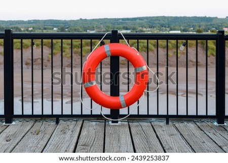 A vibrant orange polyethylene life ring hangs on a black metal railing and posts with red sand in the background. The preserver has a white grab rope. The rescue equipment is a marine buoyant throw.  Royalty-Free Stock Photo #2439253837