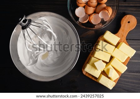 Mixing Bowl Filled with Meringue Next to Sticks of Butter: Wire whisk attachment placed in a bowl full of Swiss meringue near cracked eggs and butter Royalty-Free Stock Photo #2439257025