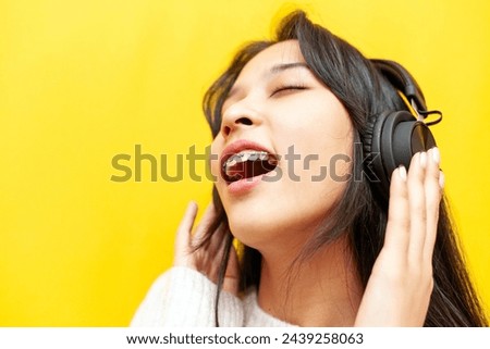 young asian woman with braces listening to music on headphones and singing on yellow isolated background, korean girl dancing to music, close-up