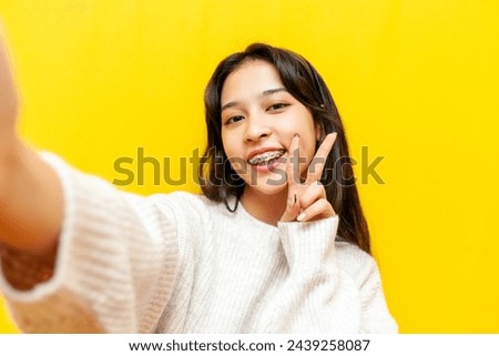 young happy asian woman with braces taking selfie and showing peace gesture on yellow isolated background, korean girl video chatting and greeting