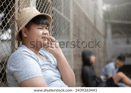 Asian teenboy in white t-shirt sits with picking teeth against a metal fence panel in a juvenile detention facility, awaiting further release, freedom and detention of people concept. Royalty-Free Stock Photo #2439263441