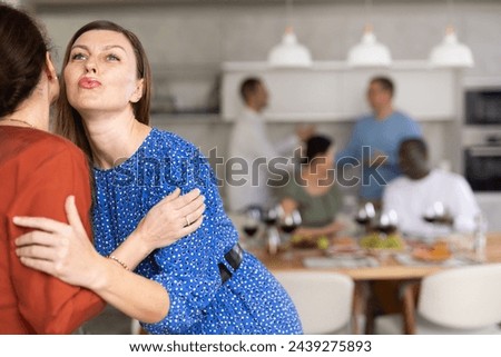 Hospitable hostess warmly greeting and kissing female friend arriving for home get-together with food and drinks on table in background.. Royalty-Free Stock Photo #2439275893