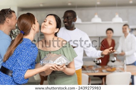 Hospitable smiling Asian woman giving greeting kiss on cheek to female friend bringing homemade dish to friendly home party Royalty-Free Stock Photo #2439275983