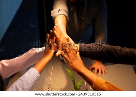 The image displays a group of hands from individuals of varying ethnicities engaging in a collective high-five above a meeting table, highlighting a moment of team success or agreement within a Royalty-Free Stock Photo #2439310515