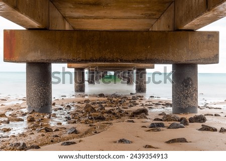 The underneath of a fishing jetty in diminishing perspective view in long exposure showing sand, rocks and support pillars on a tropical beach in Mission Beach in Queensland, Australia. Royalty-Free Stock Photo #2439354913