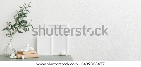 Vase with plant branches, Easter eggs, bunny and photo frames on table near white wall. Banner for design