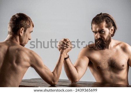 Arms wrestling thin hand, big strong arm in studio. Two man's hands clasped arm wrestling, strong and weak, unequal match. Heavily muscled bearded man arm wrestling a puny weak man.