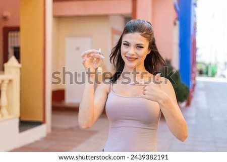 Young pretty woman holding home keys at outdoors and pointing it