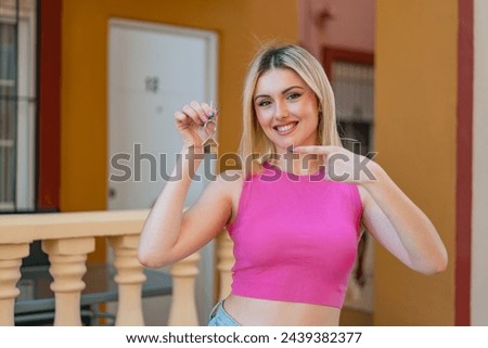Young pretty blonde woman holding home keys at outdoors and pointing it