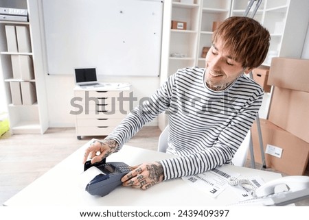 Young online store seller with credit card and payment terminal at table in warehouse