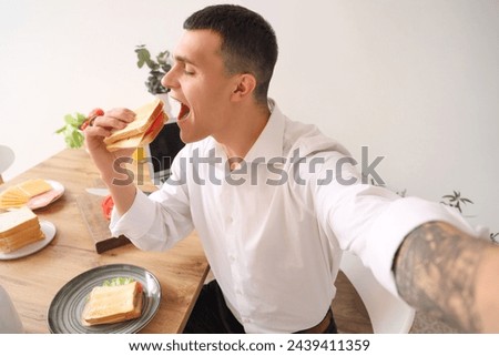 Young man taking selfie while eating crispy sandwich in kitchen