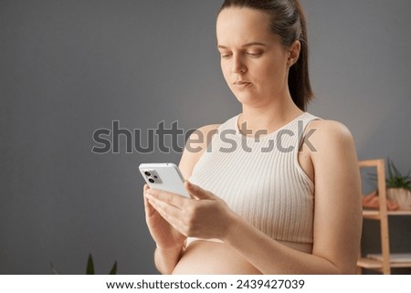 Serious brown haired pregnant woman in beige top using mobile phone browsing web pages scrolling online looking at device screen chatting with doctor gynecologist