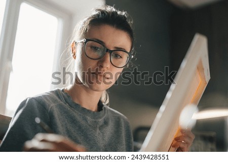 Inspired adult woman holding canvas and paintbrush, creating new artwork at home. Joyful millennial female artist drawing picture.