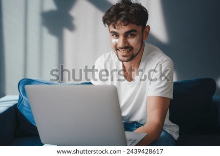 Portrait of young smiling man holding pc on lap sitting on the sofa in living room, typing on keyboard. Cheerful guy browsing internet, surfing web. People and Royalty-Free Stock Photo #2439428617