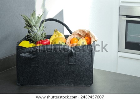 A grey eco felt bag on kitchen table with groceries, fresh vegetables and fruits. Royalty-Free Stock Photo #2439457607