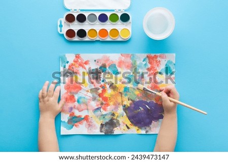 Baby boy hand holding paintbrush and drawing first colorful scratches on white paper. Light blue table background. Pastel color. Closeup. Child development. Learning painting. Point of view shot.