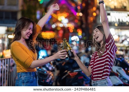 Asian woman friends hold bottle of beer, having party in front of bar. Beautiful two female traveler travel outdoor in city, enjoy spend time celebrating event on holiday vacation trip in night club.