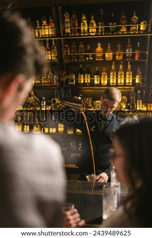Barkeeper masterfully decants alcoholic drink from shaker. Guests by bar counter supervise working barkeeper in pub with Ukrainian Trident Royalty-Free Stock Photo #2439489625
