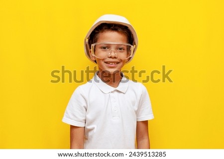 african american boy builder in hard hat and safety glasses smiling on blue isolated background, child of 10 years old foreman in uniform