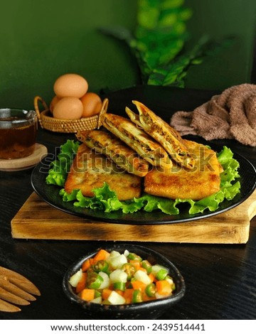 A plate of egg martabak filled with meat and vegetables with fresh henna and savory sauce with a blurry background of artificial leaves