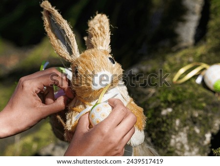 Close-up photo of hands tying a yellow ribbon around a straw Easter bunny, with a patterned Easter egg nearby, against a natural background. Ideal for concepts of Easter traditions, spring festivities