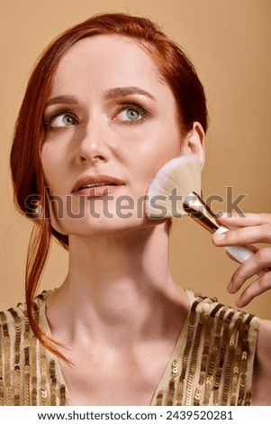 dreamy redhead woman in her 30s applying bronzer with makeup brush on beige background Royalty-Free Stock Photo #2439520281