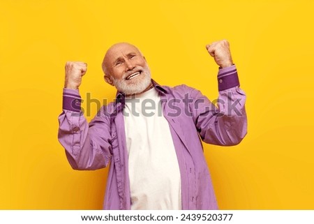 old bald grandfather in a purple shirt celebrates victory on a yellow isolated background, elderly pensioner winner rejoices at success and good luck Royalty-Free Stock Photo #2439520377